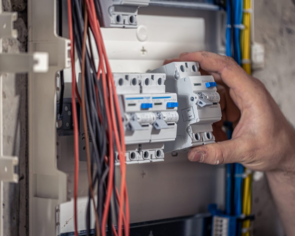 A male electrician works in a switchboard with an electrical connecting cable, connects the equipment with tools.